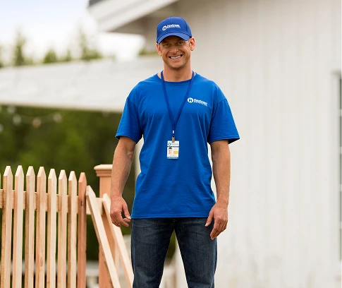 Smiling technician in a blue Handyman Connection uniform and cap stands near a wooden fence outdoors, wearing an ID badge.