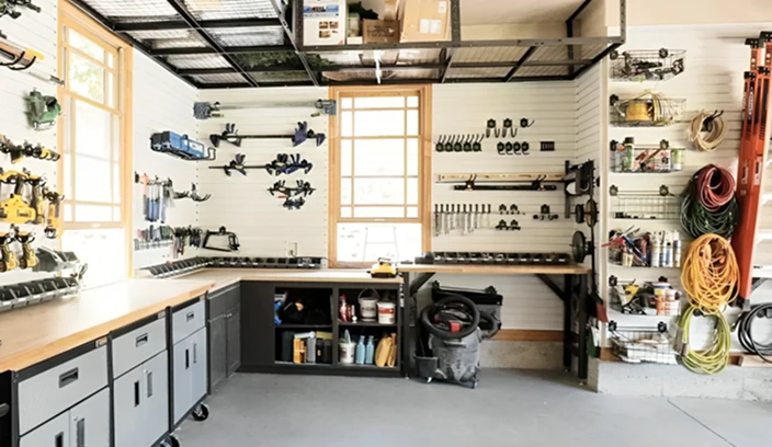 Organized garage workshop with wall-mounted tools, long workbenches, gray base cabinets, and overhead storage racks.