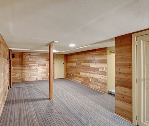 Partially finished basement with wood-paneled accent walls, carpeted floor, and support columns under recessed ceiling lights.