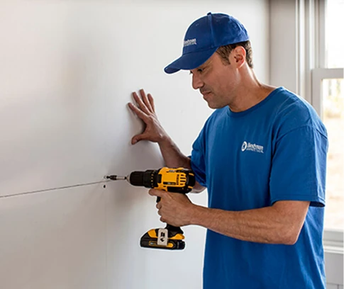 Technician in a Handyman Connection uniform uses a yellow cordless drill to drive a screw into drywall at a seam, bracing the wall with his other hand beside a window.
