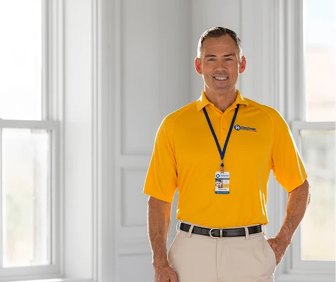 Smiling Handyman Connection home-services worker in a bright yellow polo with a lanyard and ID badge stands indoors in front of large windows.